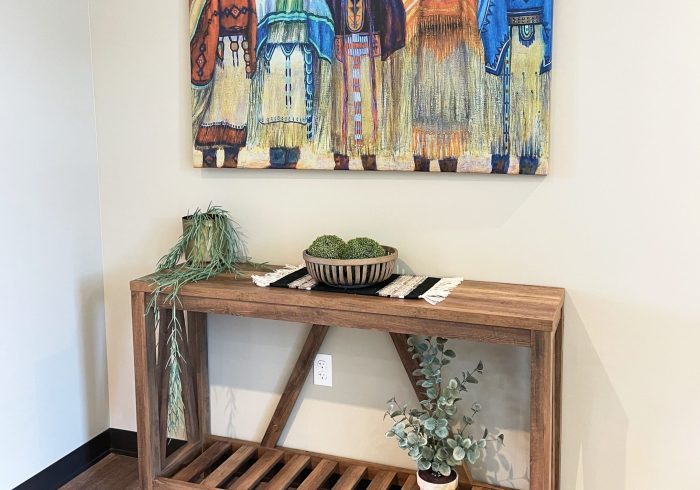 A painting of five women in regalia is hung above a wooden slatted table holding a small rug and various plants and greenery