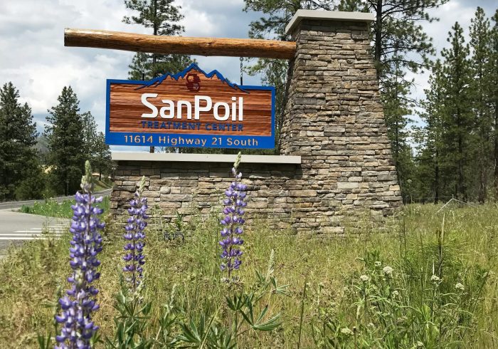 Carved wooden sign with rock base featuring name and address of San Poil Treatment center, purple and yellow flowers are blooming in the foreground