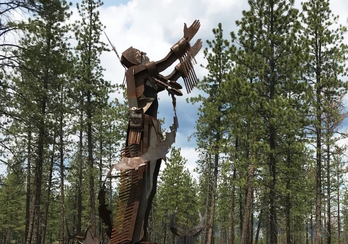 Metal sculpture of an Indigenous man in traditional clothing as seen from the side, his arms are raised toward the sky and small animals circle his feet